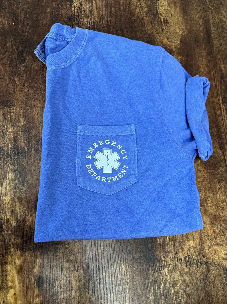 May include: A blue t-shirt with a pocket featuring the text "EMERGENCY DEPARTMENT" in white. The pocket design includes a white medical symbol. The shirt is folded on a wooden surface.