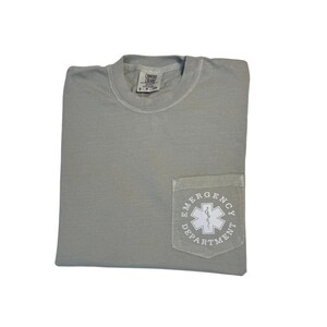 May include: A sage green t-shirt with a pocket featuring a white emergency medical services (EMS) symbol and the words "Emergency Department." The shirt is folded and has a tag at the neckline.