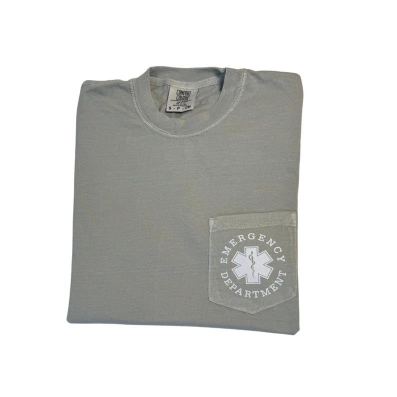 May include: A sage green t-shirt with a pocket featuring a white emergency medical services (EMS) symbol and the words "Emergency Department." The shirt is folded and has a tag at the neckline.