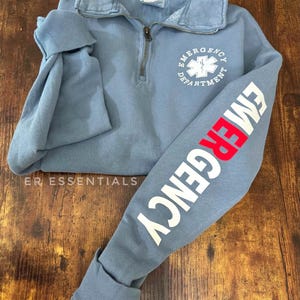 May include: A light blue sweatshirt with a white "Emergency Department" logo featuring a red star of life on the front and a white "Emergency" graphic on the left sleeve.