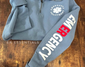 Op de afbeelding: Een lichtblauw sweatshirt met een wit "Emergency Department" logo met een rode ster van het leven op de voorkant en een witte "Emergency" afbeelding op de linkermouw.