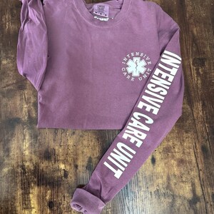 May include: A mauve long-sleeve shirt with the words "INTENSIVE CARE UNIT" printed in white on the sleeve. The shirt features a white medical symbol and the words "INTENSIVE CARE UNIT" on the front.