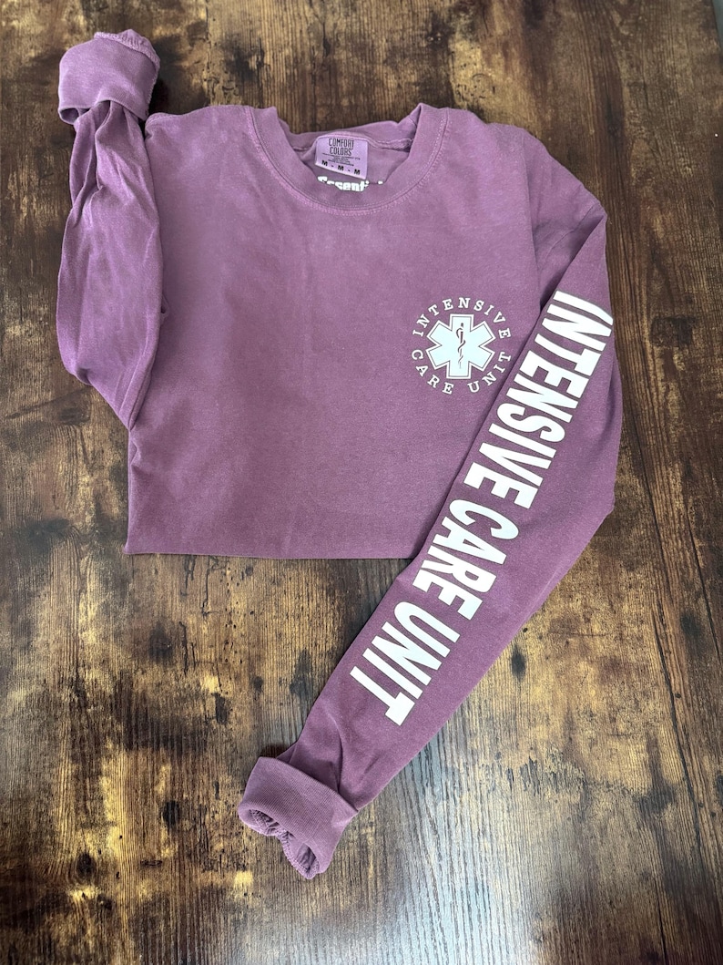 May include: A mauve long-sleeve shirt with the words "INTENSIVE CARE UNIT" printed in white on the sleeve. The shirt features a white medical symbol and the words "INTENSIVE CARE UNIT" on the front.