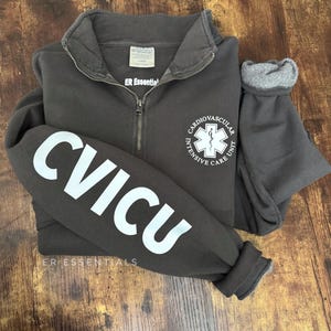 May include: A dark gray zippered sweatshirt with the text "CVICU" printed in white on the left sleeve. The right side of the sweatshirt has a white logo with a star of life and the text "Cardiovascular Intensive Care Unit". The text "ER Essentials" is printed in white below the logo.