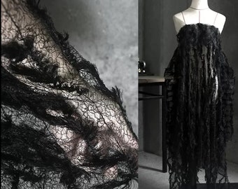 Puede incluir: Tela de encaje negra con un borde deshilachado. La tela es delicada y tiene un tejido suelto. Se muestra drapeada sobre una superficie y en un maniquí.