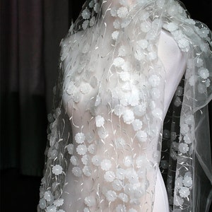 May include: White floral lace fabric with 3D flowers. The fabric is sheer and delicate, with a soft, flowing texture. The flowers are small and delicate, with a slightly raised design.
