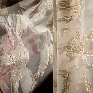 Puede incluir: Primer plano de una tela transparente con un diseño fluido y orgánico. La tela es principalmente blanquecina con acentos bordados de color dorado. La textura es delicada y translúcida, con un brillo sutil. El diseño parece ser un paisaje estilizado.