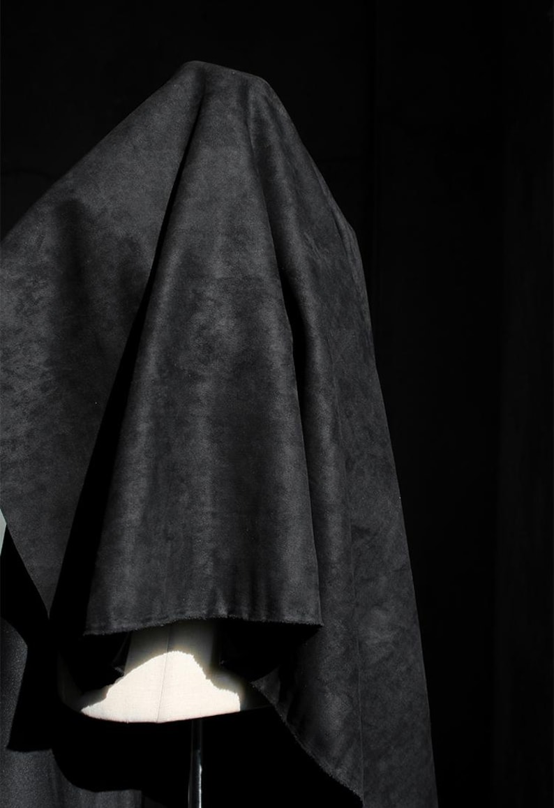 May include: A draped, dark gray fabric with a textured appearance. The fabric is draped over a white mannequin form, creating folds and shadows. The background is a solid black.