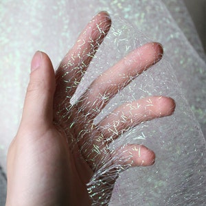 May include: A hand holding a piece of iridescent fabric. The fabric is a sheer, open weave with a shimmering, glittery effect. The colour shifts between white, silver, and hints of green and blue. The fabric appears delicate and lightweight.