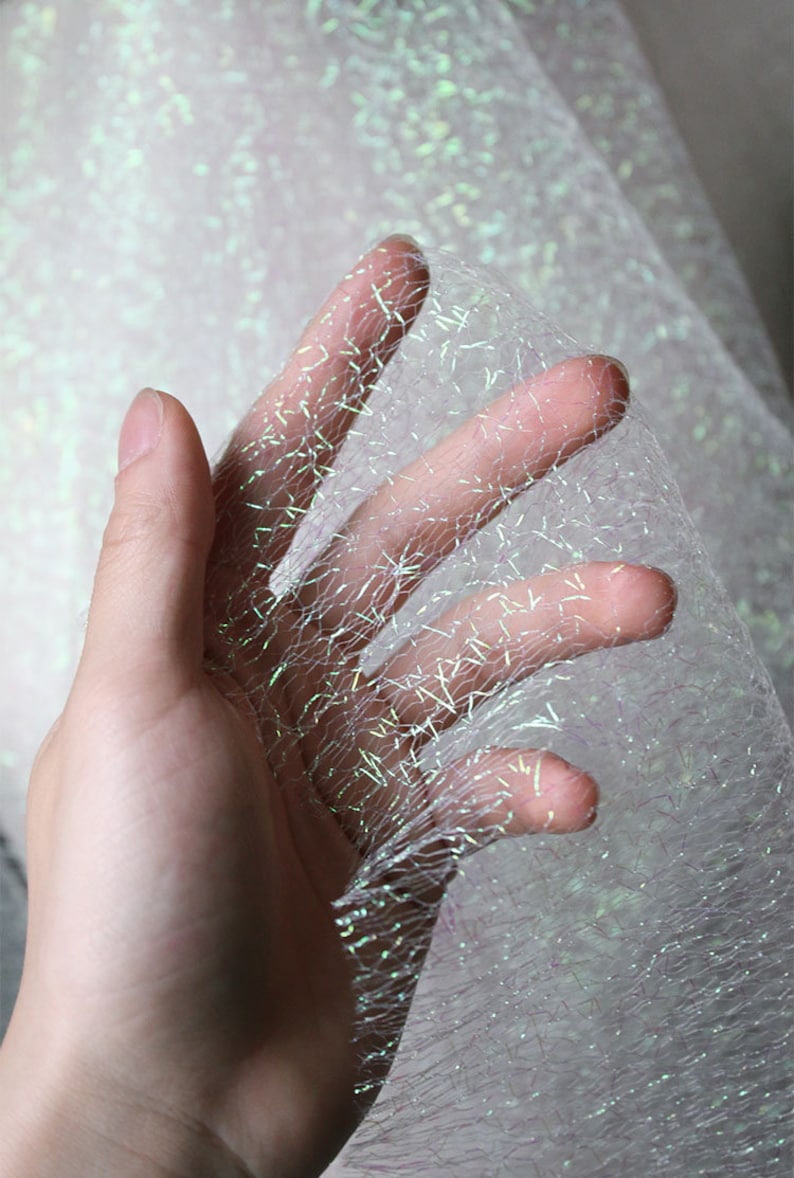 May include: A hand holding a piece of iridescent fabric. The fabric is a sheer, open weave with a shimmering, glittery effect. The colour shifts between white, silver, and hints of green and blue. The fabric appears delicate and lightweight.