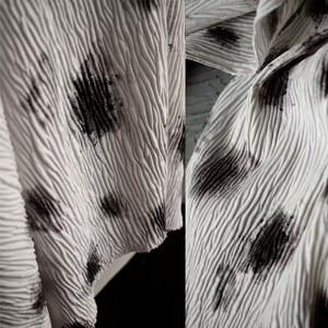 May include: Close-up of a white, textured fabric with a rippled surface. The fabric is adorned with irregular black markings, creating a spotted pattern. The material appears to be draped, showcasing its texture and design.