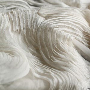 Op de afbeelding: Witte stof met een textuur, gerimpeld en gelaagd ontwerp. De stof heeft een zachte, vloeiende uitstraling.
