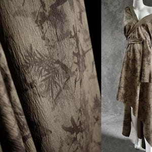 May include: A draped, olive-green fabric with a textured surface and a dark brown floral pattern. The fabric is used to create a wrap dress with a matching belt, displayed on a mannequin.