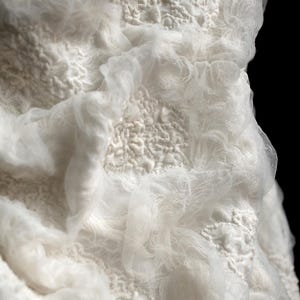 May include: Close-up of a white textile with a layered design. The fabric has a textured, almost bubbly appearance, with areas of raised, intricate patterns. Sheer, flowing material drapes over the textured base, creating depth and dimension.