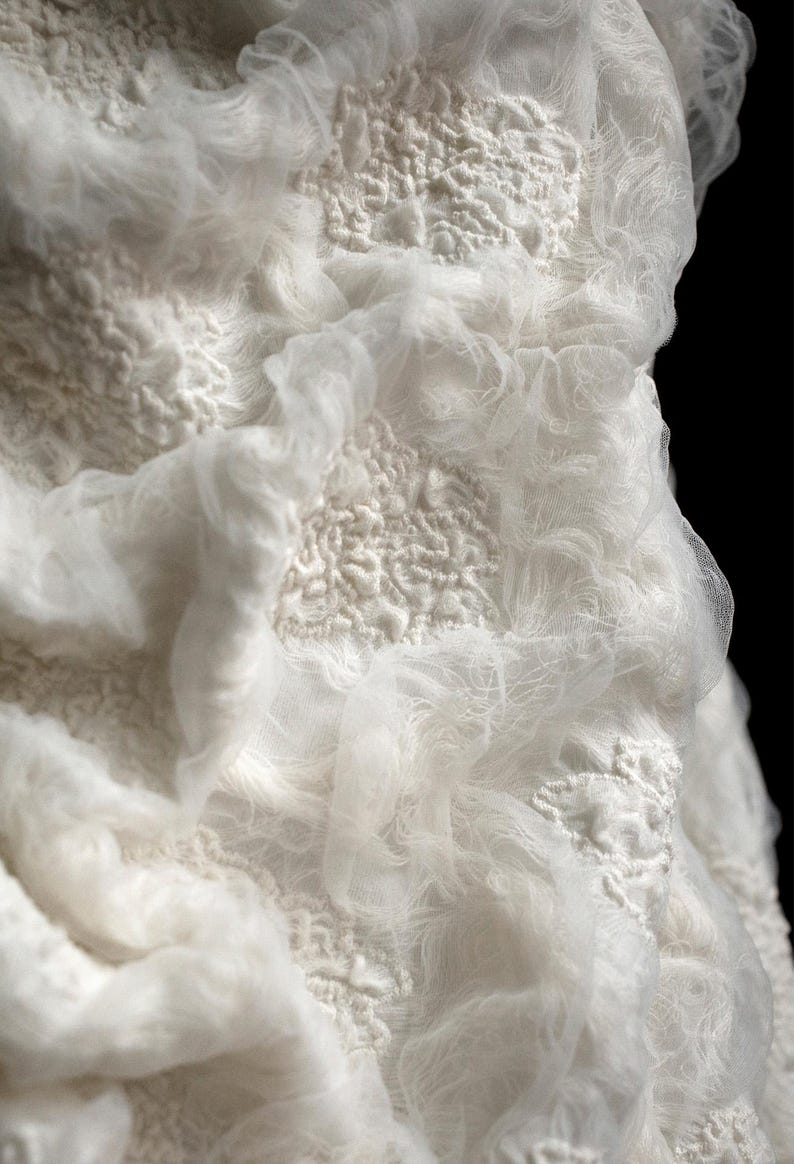 May include: Close-up of a white textile with a layered design. The fabric has a textured, almost bubbly appearance, with areas of raised, intricate patterns. Sheer, flowing material drapes over the textured base, creating depth and dimension.