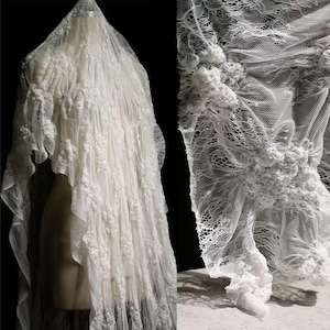 Op de afbeelding: Een witte kanten sluier met ingewikkelde bloemen- en textuurdetails. De sluier is over een mannequin gedrapeerd en toont het delicate ontwerp en de vloeiende stof. Een close-up toont het gedetailleerde kantpatroon.