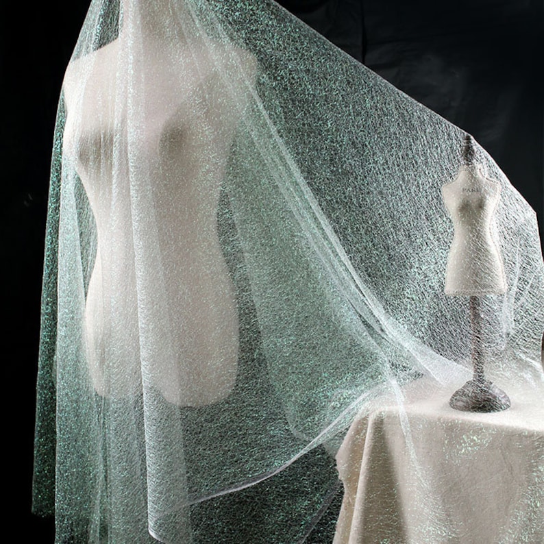 May include: A light, shimmering fabric draped over a mannequin. The fabric has a delicate, textured appearance with a subtle sparkle. A smaller mannequin is on a table in the background. The fabric is a light, ethereal colour.