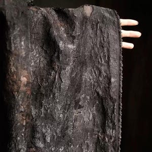May include: A close-up of a textured, black fabric with a rough, uneven surface. The fabric appears to be draped, with visible folds and a slightly shimmering effect. The edges of the fabric are frayed, adding to its unique texture.