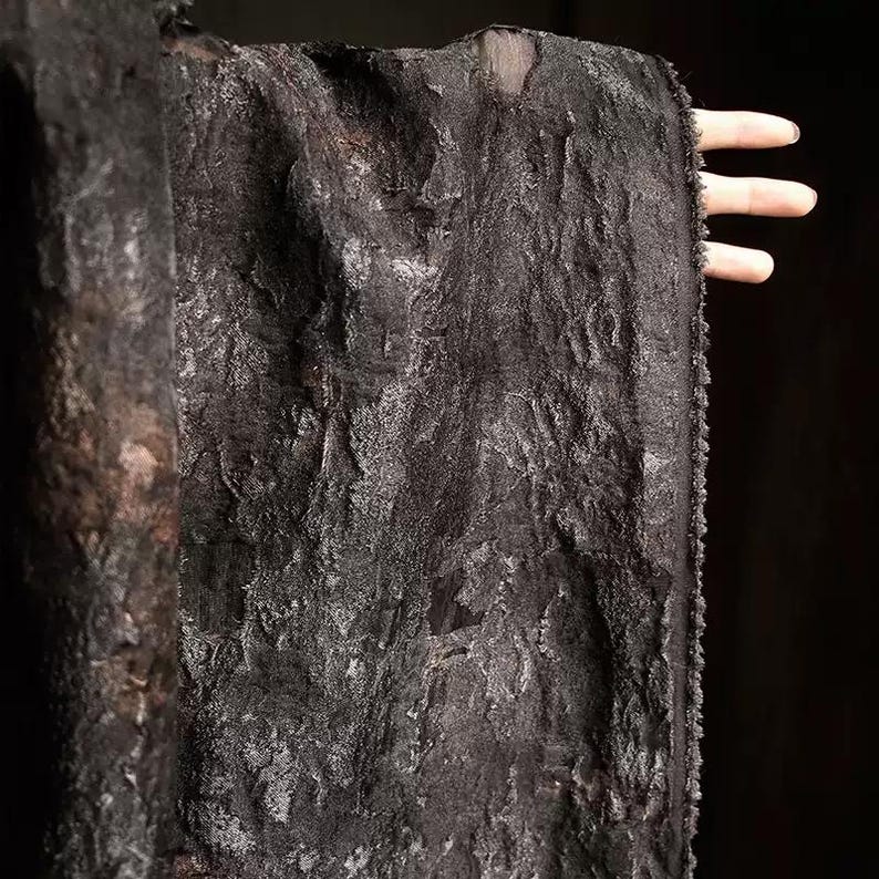 May include: A close-up of a textured, black fabric with a rough, uneven surface. The fabric appears to be draped, with visible folds and a slightly shimmering effect. The edges of the fabric are frayed, adding to its unique texture.