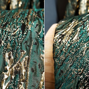 May include: Close-up of a green and gold fabric with a textured, embossed pattern. The fabric has a metallic sheen and a subtle shimmer.