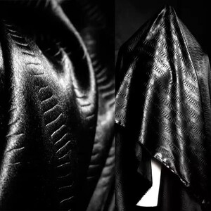 May include: Black fabric with a textured, embossed pattern. The fabric is draped over a surface, creating folds and shadows.