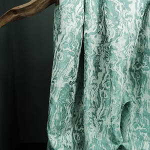 Peut inclure: Un tissu drapé avec un motif marbré dans des tons de vert d'eau et de blanc. Le tissu est suspendu à une branche en bois, créant des plis et des ombres. L'ensemble est élégant et texturé, idéal pour la décoration intérieure.