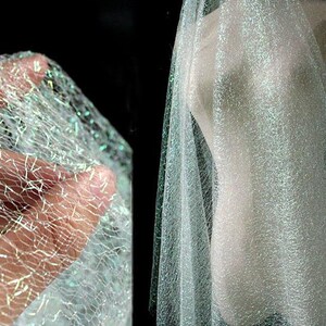 May include: A close-up shot of a shimmering, iridescent fabric with a net-like design. The fabric has a delicate, airy appearance, with a subtle sheen that catches the light. The color is a soft, pale white with hints of green and blue.