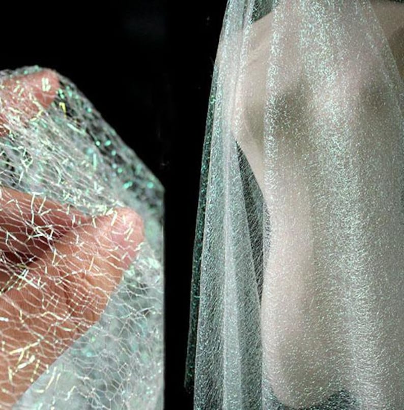 May include: A close-up shot of a shimmering, iridescent fabric with a net-like design. The fabric has a delicate, airy appearance, with a subtle sheen that catches the light. The color is a soft, pale white with hints of green and blue.