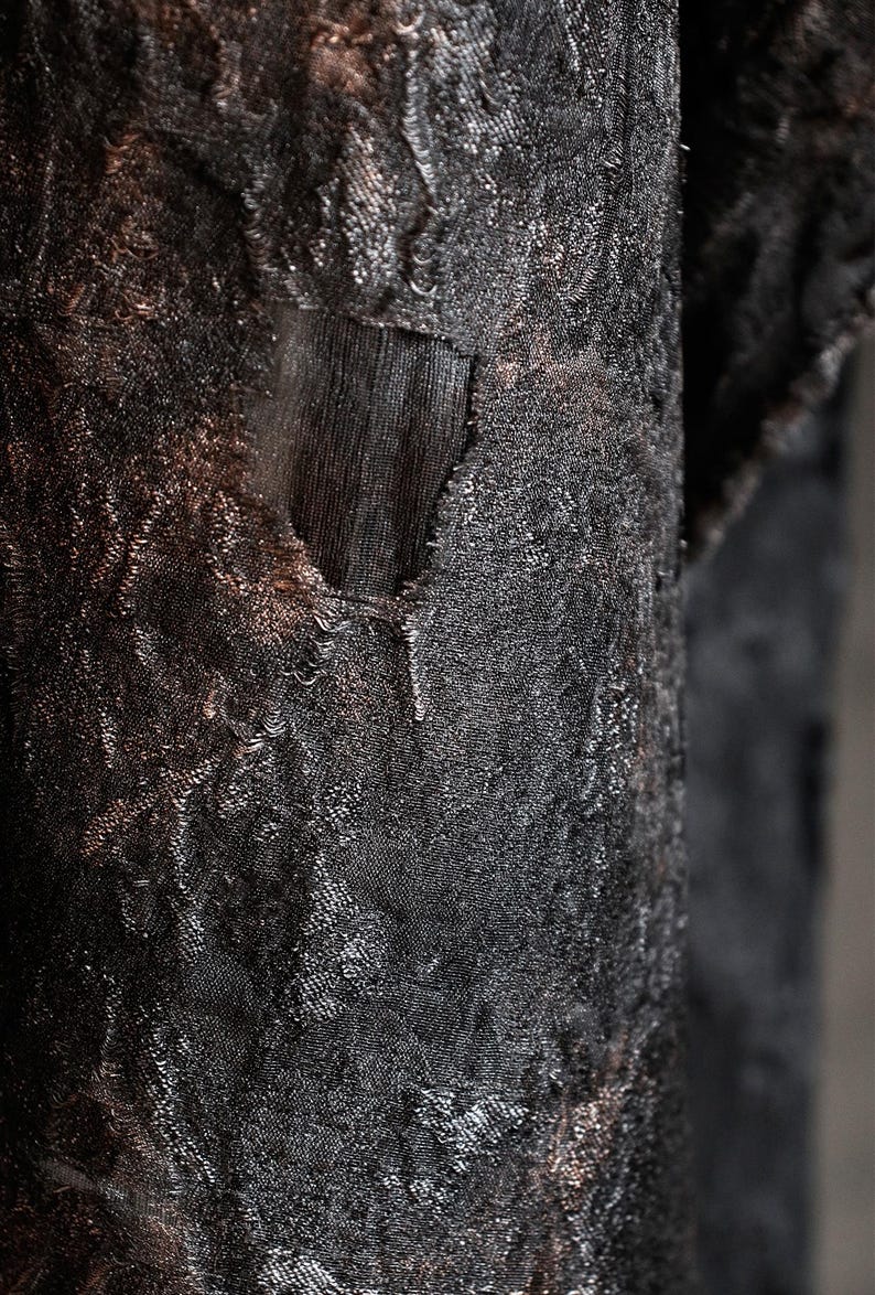 May include: Close-up of a textured, dark fabric with a distressed appearance. The fabric has a complex pattern and a torn section, revealing a darker layer beneath. The overall color is a deep black with hints of brown.