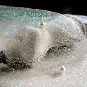 May include: A close-up shot of a white, net-like fabric with iridescent threads, creating a subtle shimmer. Two faux pearls are scattered across the surface. A small pouch with the word "PARIS" is visible, adding a touch of elegance to the scene.