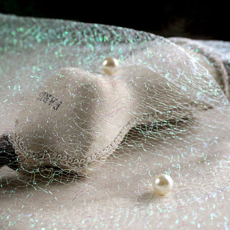 May include: A close-up shot of a white, net-like fabric with iridescent threads, creating a subtle shimmer. Two faux pearls are scattered across the surface. A small pouch with the word "PARIS" is visible, adding a touch of elegance to the scene.