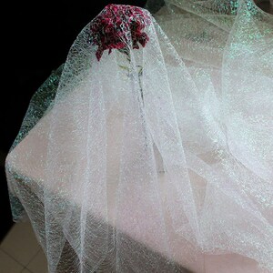 May include: A red flower is partially veiled by a sheer, white, iridescent fabric. The fabric is draped over a pale pink surface, creating a soft, ethereal effect. The dark background highlights the delicate texture of the fabric.