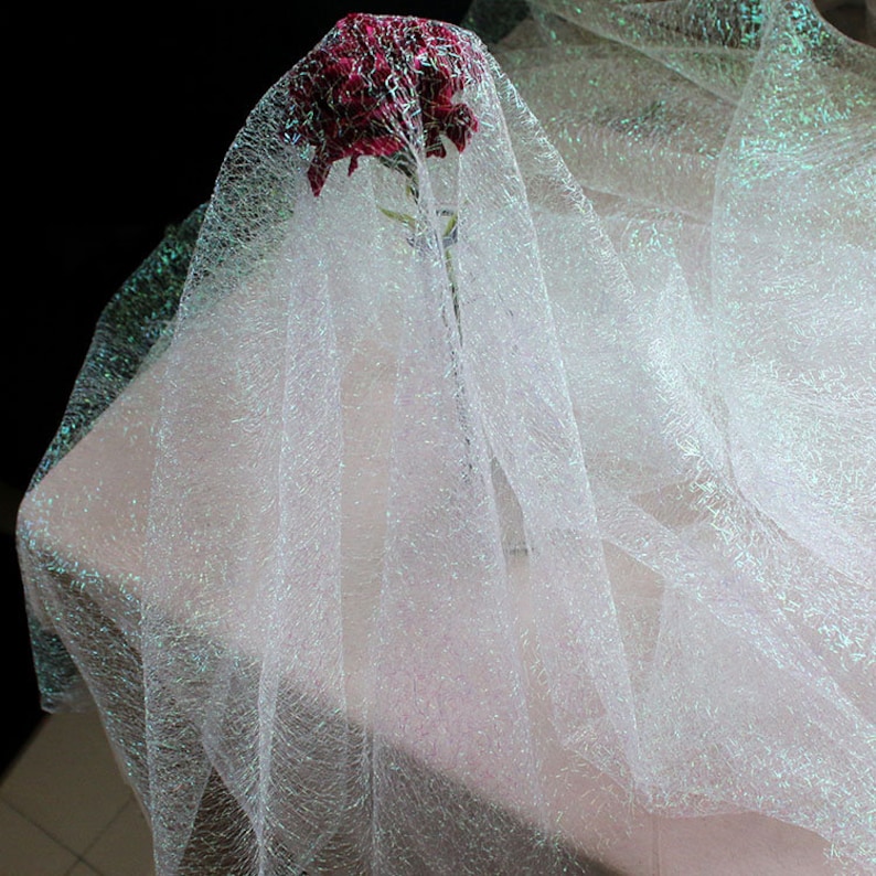 May include: A red flower is partially veiled by a sheer, white, iridescent fabric. The fabric is draped over a pale pink surface, creating a soft, ethereal effect. The dark background highlights the delicate texture of the fabric.