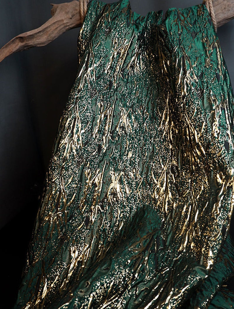 May include: Green fabric with a gold metallic pattern. The fabric has a textured, woven look.