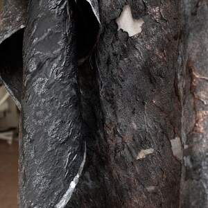 May include: Close-up of textured, dark fabric with a layered appearance. The material has a rough, uneven surface with a mix of black, gray, and brown tones. The fabric appears to be draped or folded, revealing glimpses of a lighter, neutral-colored backing.