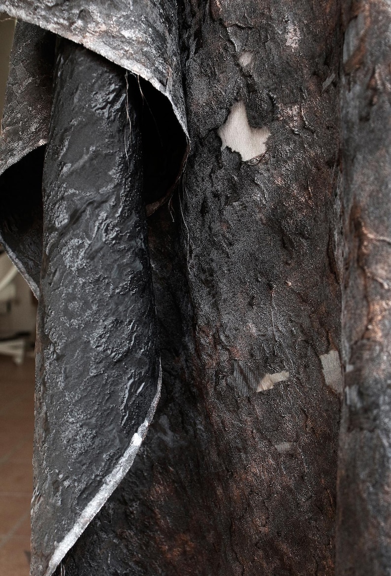 May include: Close-up of textured, dark fabric with a layered appearance. The material has a rough, uneven surface with a mix of black, gray, and brown tones. The fabric appears to be draped or folded, revealing glimpses of a lighter, neutral-colored backing.
