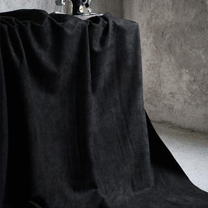 May include: A vintage black sewing machine sits atop a draped black fabric. The fabric is heavily draped, creating deep shadows and folds. The background is a textured gray wall.