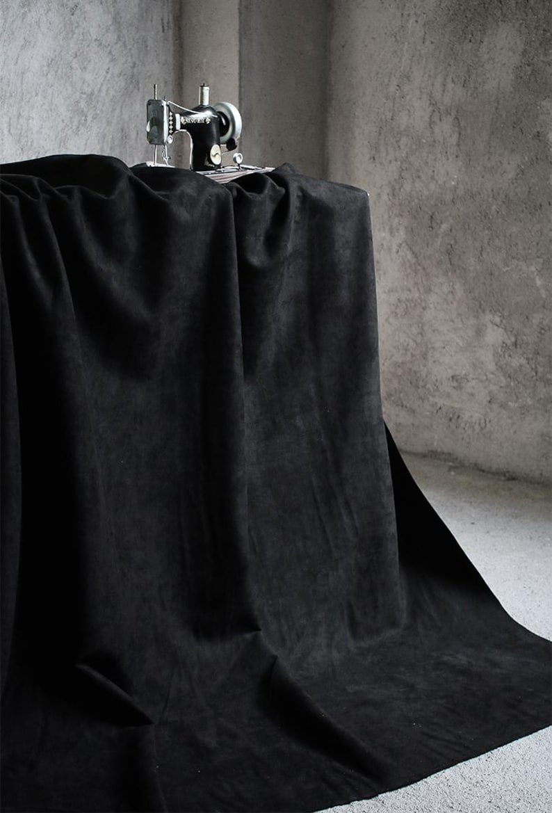 May include: A vintage black sewing machine sits atop a draped black fabric. The fabric is heavily draped, creating deep shadows and folds. The background is a textured gray wall.
