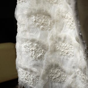 May include: Close-up of a white, textured fabric with raised floral-like patterns. The fabric appears to be layered, with a sheer, gauzy material over a more solid base. The edges are frayed, adding to the handmade aesthetic.