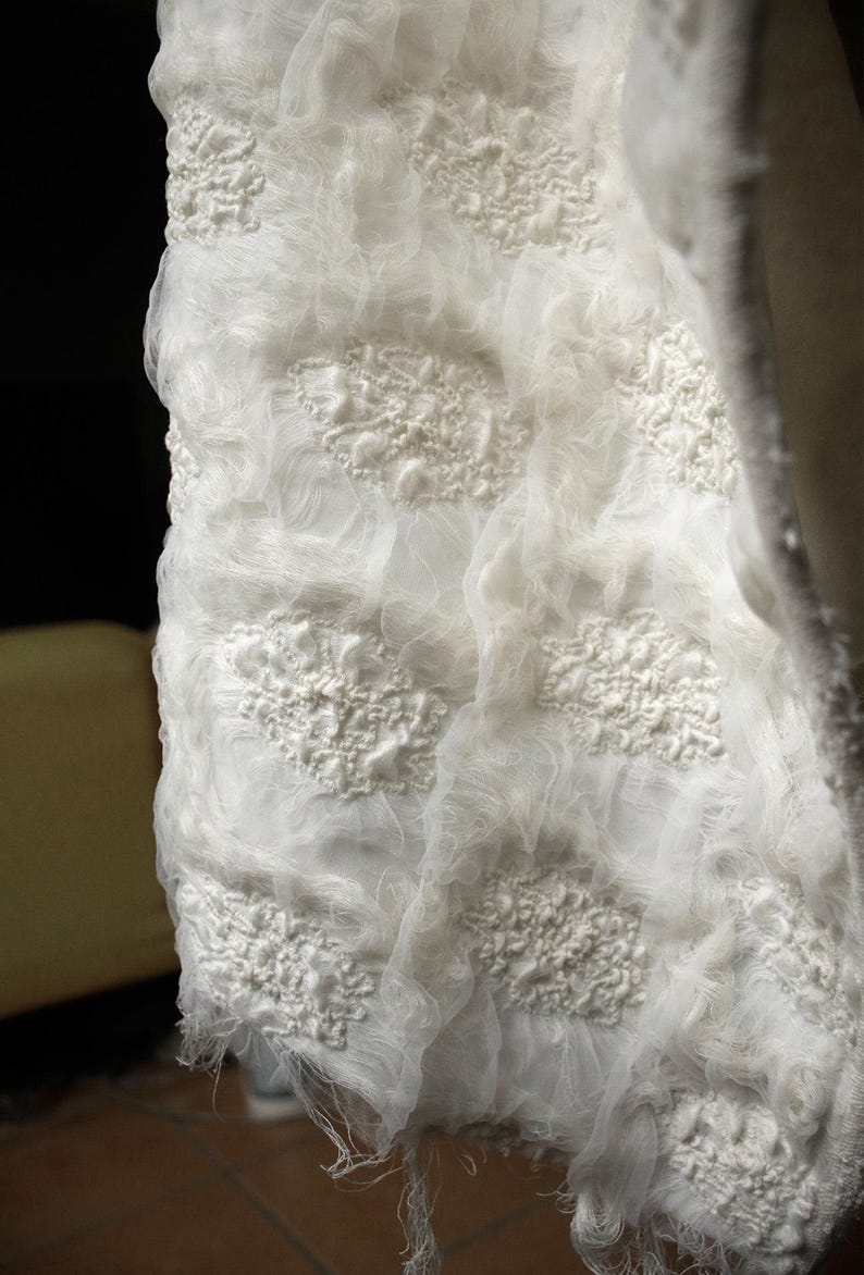 May include: Close-up of a white, textured fabric with raised floral-like patterns. The fabric appears to be layered, with a sheer, gauzy material over a more solid base. The edges are frayed, adding to the handmade aesthetic.