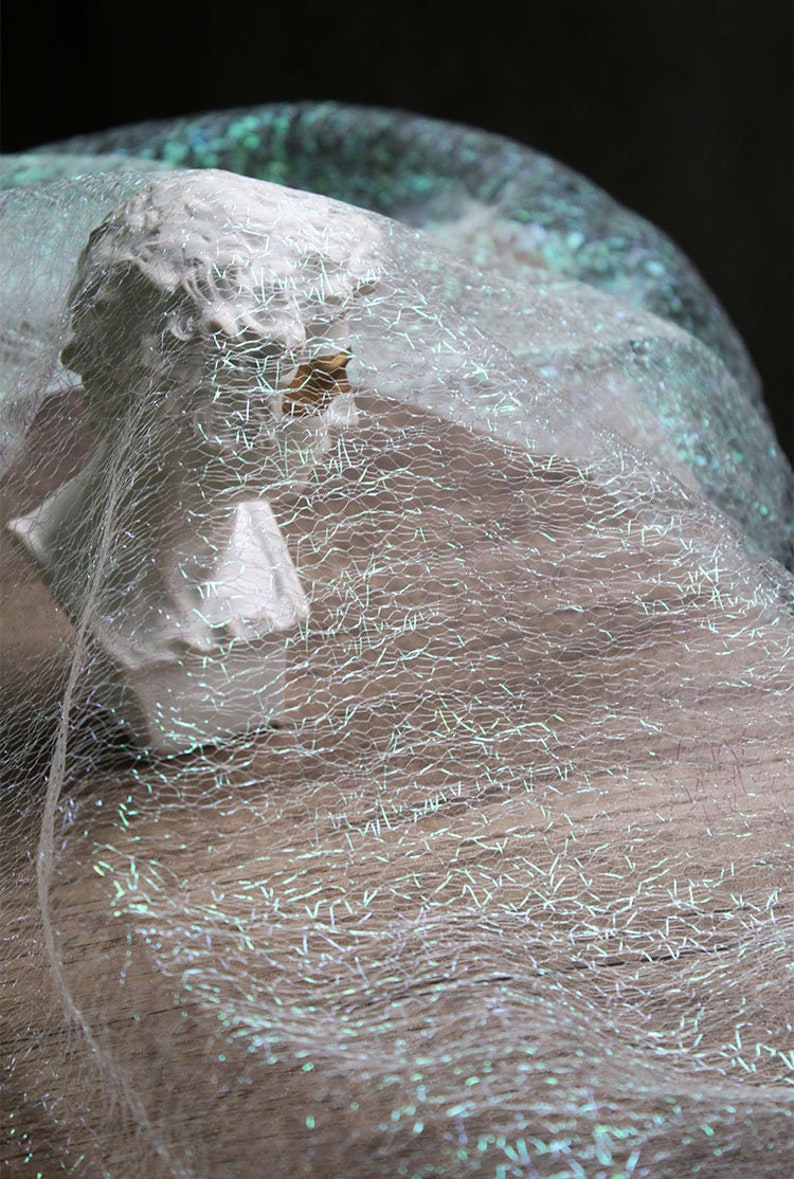 May include: A small, white bust of a classical figure is partially covered by a shimmering, iridescent fabric. The fabric has a delicate, web-like pattern and reflects light with a subtle, ethereal glow. The bust is set against a dark background.
