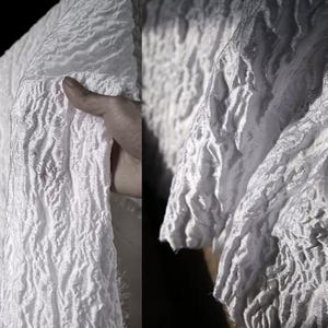 Puede incluir: Primer plano de tela blanca con superficie texturizada y arrugada. La tela está sostenida por una mano, revelando la profundidad y el patrón del material. La tela parece ser un material ligero, posiblemente para ropa o decoración del hogar.