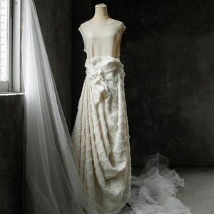 May include: A cream-colored wedding dress on a mannequin, featuring a draped skirt and a textured bodice. A long, sheer veil hangs near a window, with soft light illuminating the fabric. The dress is displayed in a dimly lit room.