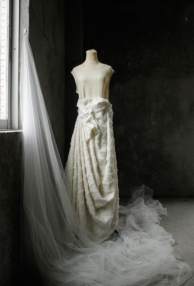 May include: A cream-colored wedding dress on a mannequin, featuring a draped skirt and a textured bodice. A long, sheer veil hangs near a window, with soft light illuminating the fabric. The dress is displayed in a dimly lit room.