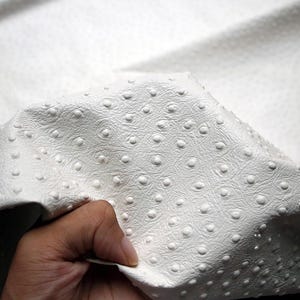 Pode incluir: Tecido de couro sintético branco com um padrão texturizado e estampado de pequenos círculos.