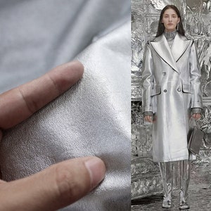 Può includere: Una giacca di pelle argentata con chiusura a doppio petto e un ampio colletto. La giacca è mostrata su una modella in piedi in una stanza argentata.