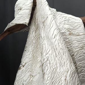 May include: White fabric with a textured, wavy pattern. The fabric has a subtle sheen and is draped over a wooden surface.