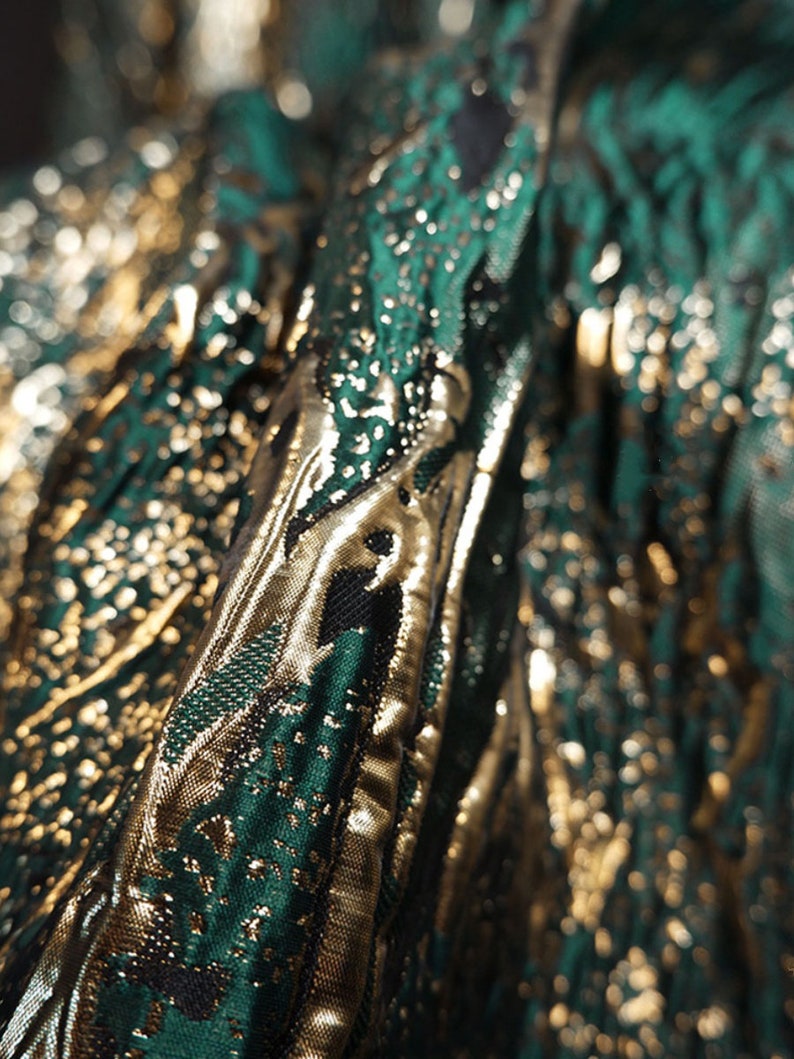 May include: Close-up of a green and gold fabric with a textured pattern. The fabric has a metallic sheen and is likely used for clothing or upholstery.