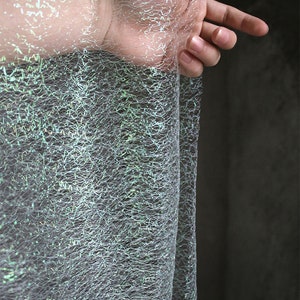 May include: A close-up of a shimmering, web-like fabric in shades of grey and silver, with hints of green and blue. The fabric has an iridescent quality, and is draped. A hand is visible, interacting with the material.