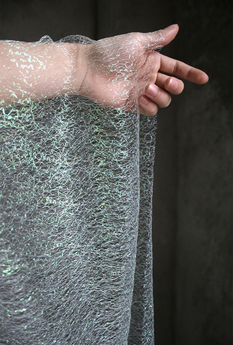 May include: A close-up of a shimmering, web-like fabric in shades of grey and silver, with hints of green and blue. The fabric has an iridescent quality, and is draped. A hand is visible, interacting with the material.